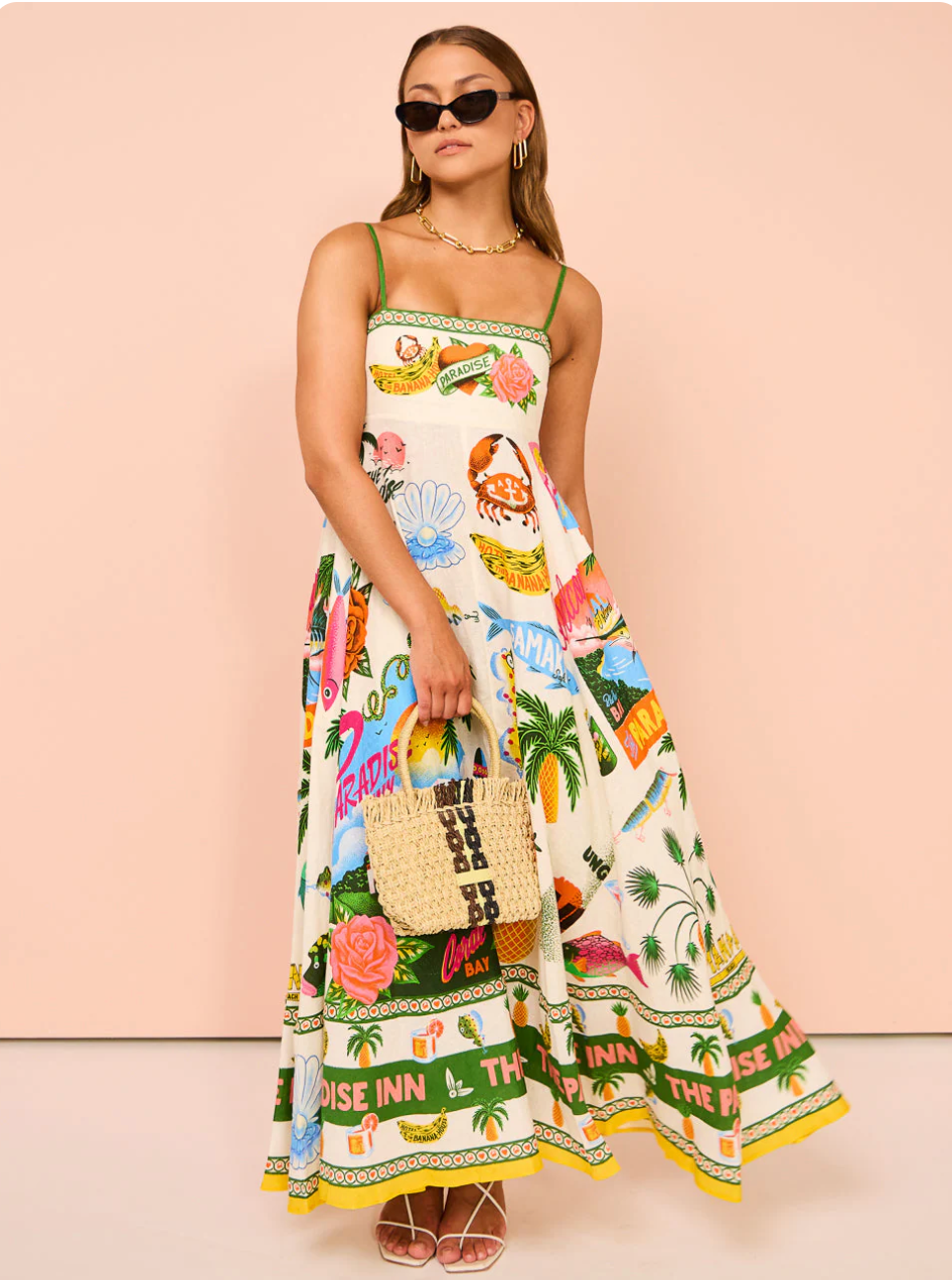 Women's smocked maxi dress with vibrant tropical print, green straps, and a yellow border, paired with a straw bag.