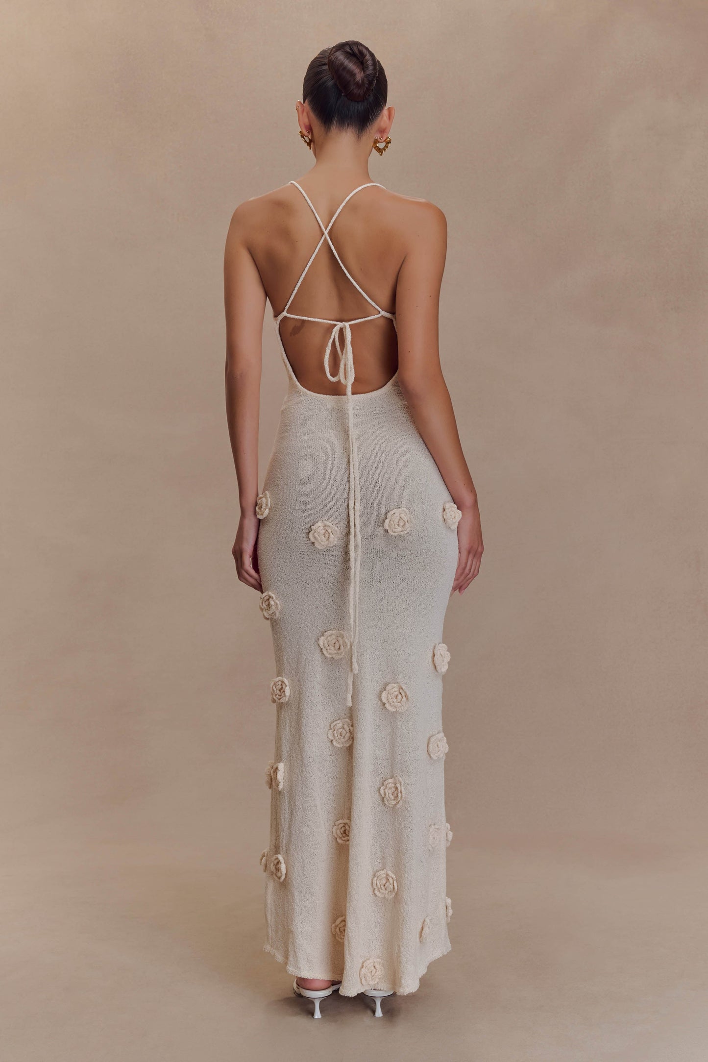 Back view of a cream knit maxi dress featuring criss-cross spaghetti straps and a low open back, adorned with 3D floral details.