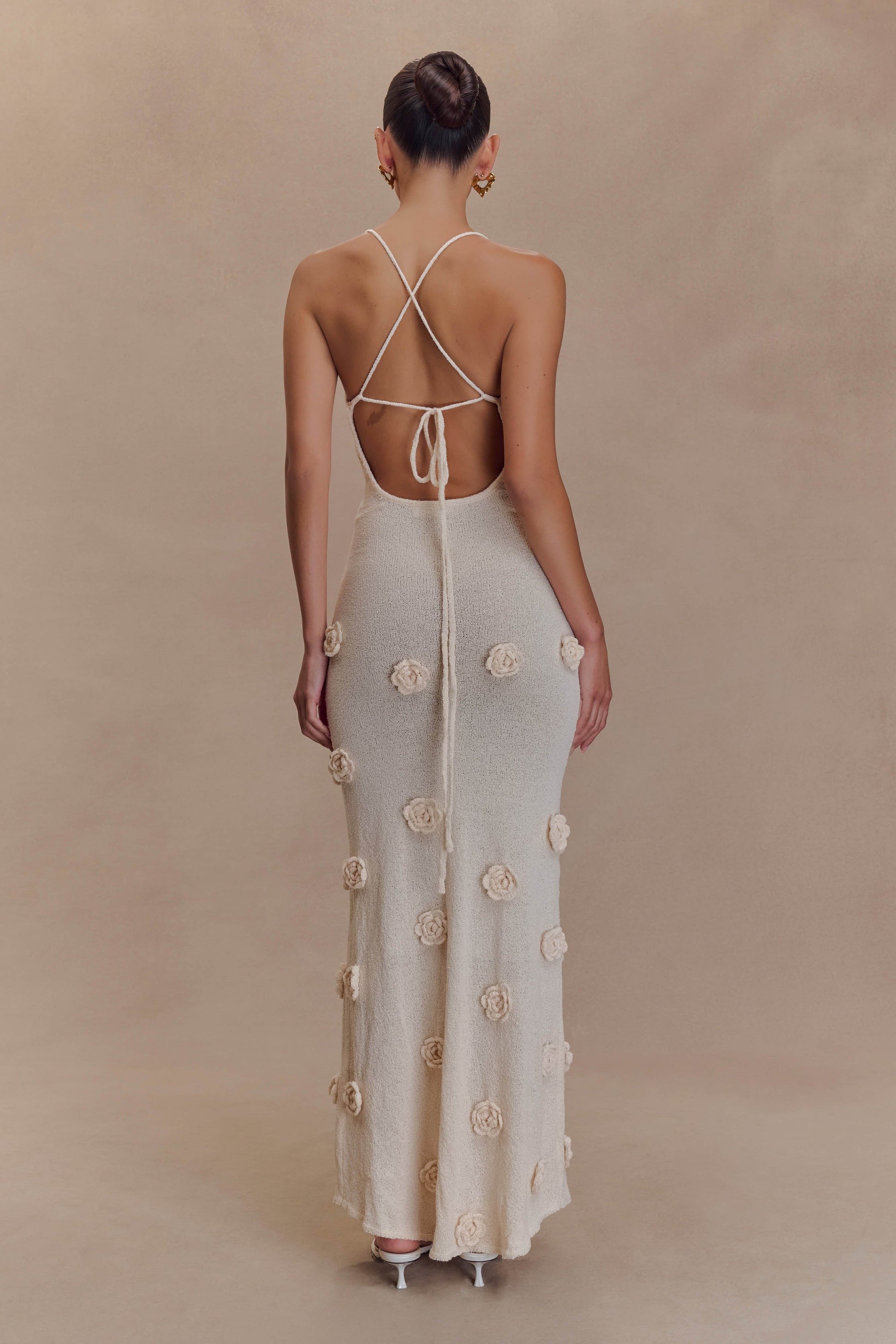 Back view of a cream knit maxi dress featuring criss-cross spaghetti straps and a low open back, adorned with 3D floral details.