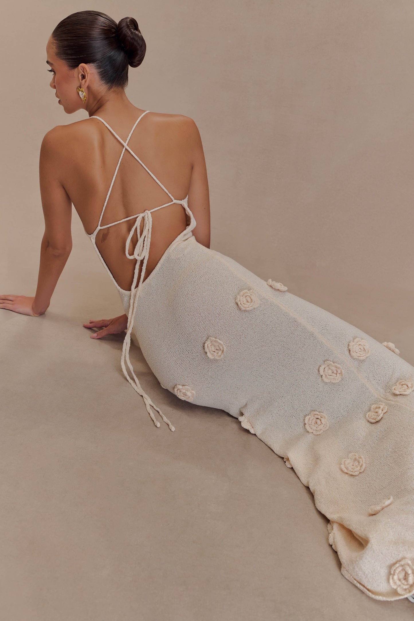 Side profile of a woman sitting, showcasing the cream knit maxi dress with 3D floral appliques and a low open back with criss-cross straps.
