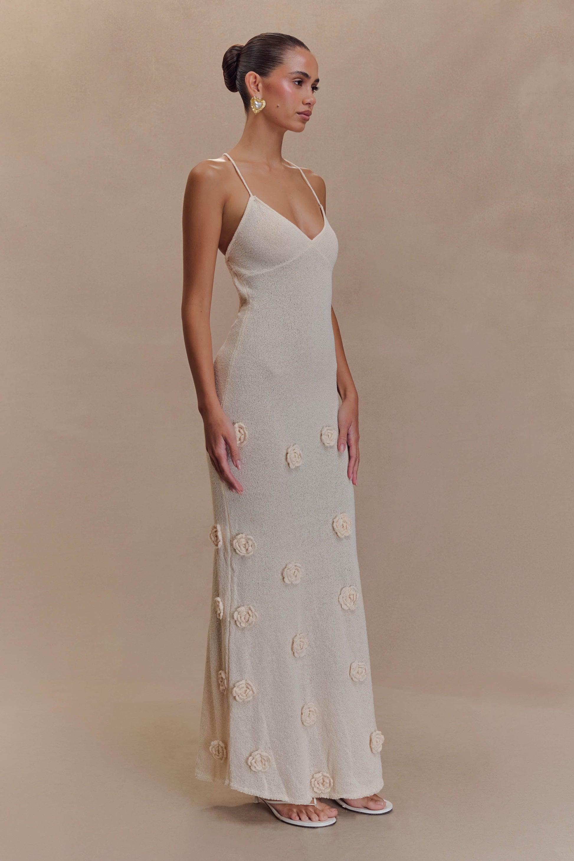 Side view of a woman standing in a cream knit maxi dress with a v-neckline, spaghetti straps, and scattered 3D floral appliques.