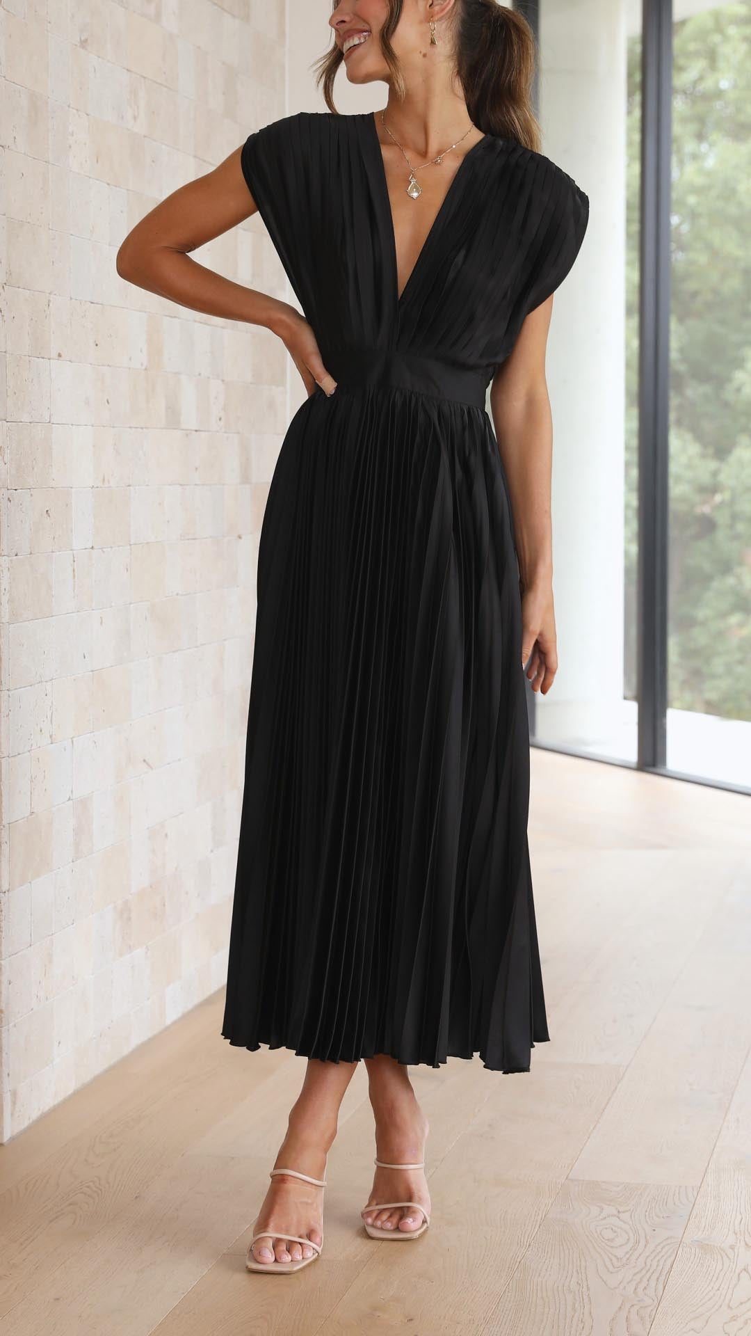 Black pleated midi dress with deep v-neckline and cap sleeves, posed with hand on hip, a chic wedding guest dress.