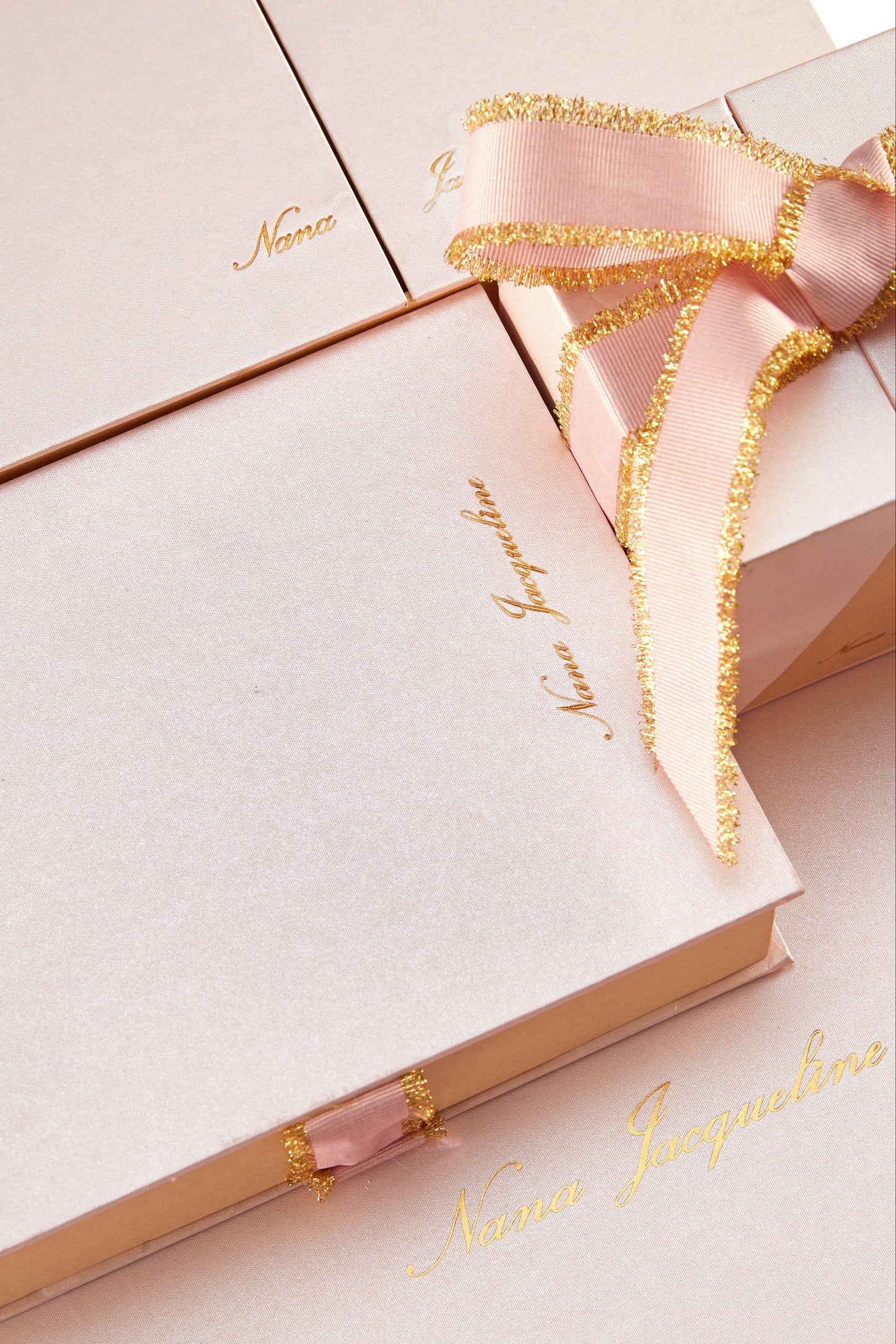 Close-up of a stack of light pink gift boxes with gold embossed lettering and a pink and gold ribbon, suggesting a luxurious presentation for the dress.