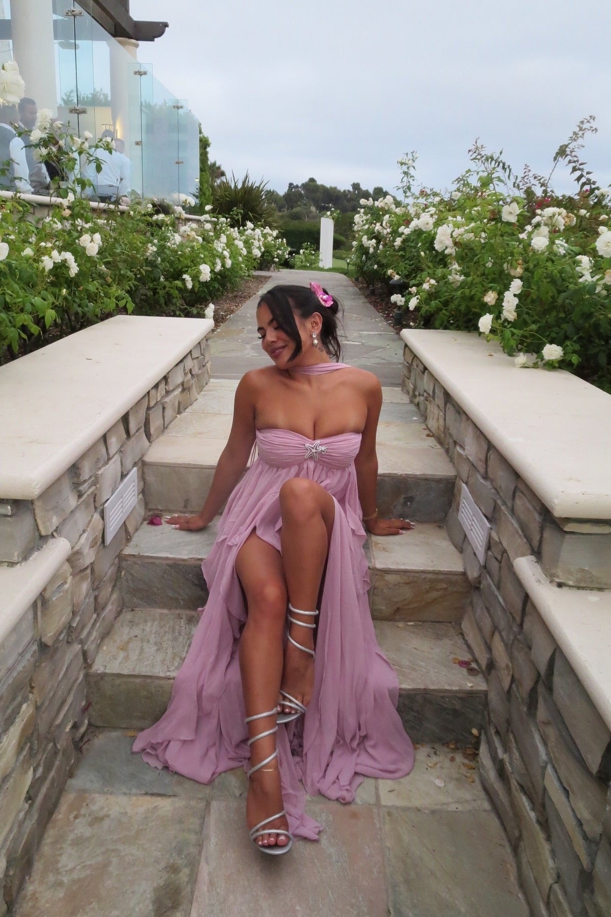 Woman sitting on stone steps in a light pink strapless maxi dress with a high slit and ruffled skirt, showcasing silver strappy heels, a great wedding guest dress.