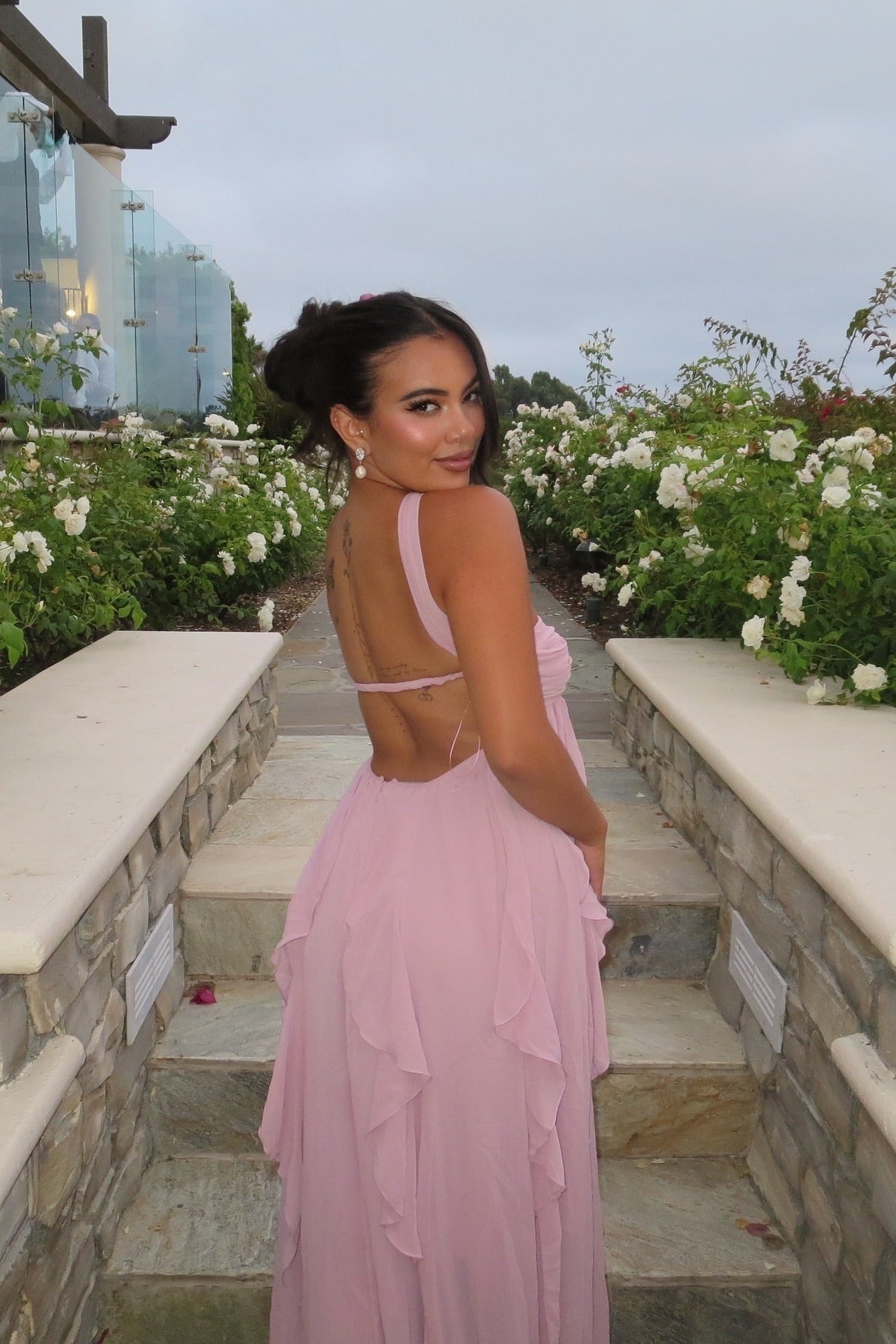 Back view of a woman on stone steps, looking over her shoulder, wearing a light pink strapless maxi dress with a low back and ruffled skirt, suitable for wedding guests.